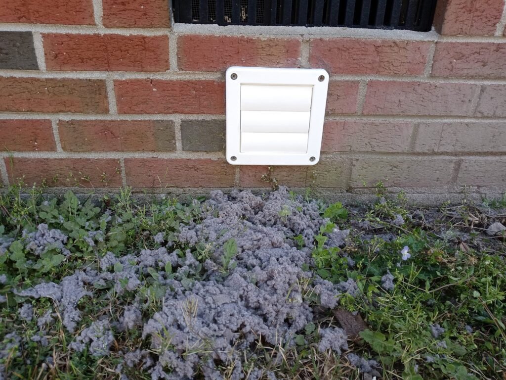 The same exterior dryer vent after cleaning, with a new white louvered cover fitted and the removed lint piled on the ground below
